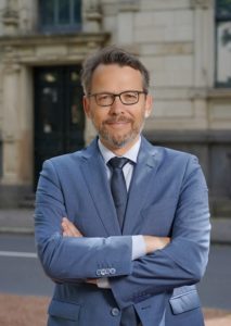 Otto Fricke im blauen Anzug steht mit verschränkten Armen vor einem historischen Gebäude und lächelt in die Kamera.